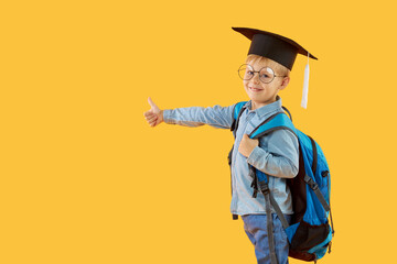 Child boy primary school student in a graduate cap, glasses and with a backpack on a yellow background. Shows ok sign with his finger. Knowledge day and back to school. Copy space