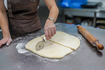 Young female baker cutting dough on table with pizza cutter
