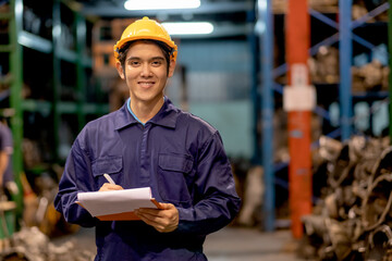 Asian factory man or warehouse worker with smiling hold clipboard and write data of product quality also look to camera in automotive parts workplace area. He looks happy with working in industrial.