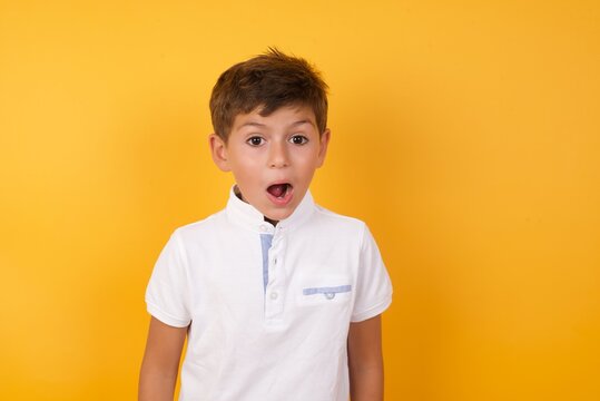 little cute Caucasian boy kid wearing white t-shirt against yellow wall having stunned and shocked look, with mouth open and jaw dropped exclaiming: Wow, I can't believe this. Surprise and shock