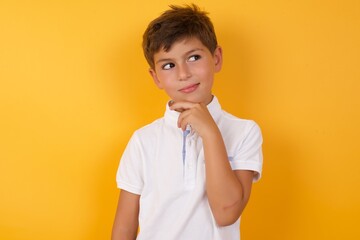 Dreamy little cute Caucasian boy kid wearing white t-shirt against yellow wall with pleasant...