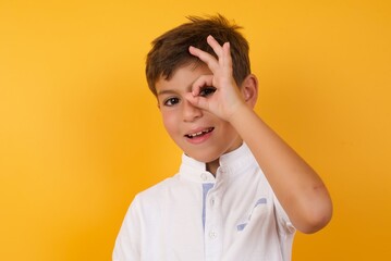 little cute Caucasian boy kid wearing white t-shirt against yellow wall with happy face smiling doing ok sign with hand on eye looking through finger.