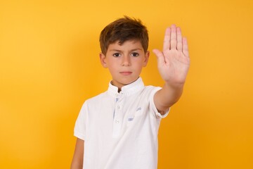 little cute Caucasian boy kid wearing white t-shirt against yellow wall doing stop gesture with palm of the hand. Warning expression with negative and serious gesture on the face.