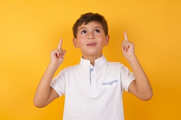 Successful friendly looking little cute Caucasian boy kid wearing white t-shirt against yellow wall exclaiming excitedly, pointing both index fingers up, indicating something.