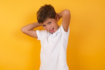 little cute boy kid wearing white t-shirt against yellow wall relaxing and stretching, arms and hands behind head and neck smiling happy