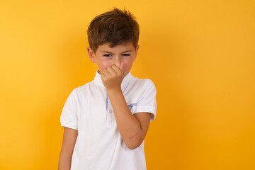 little cute boy kid wearing white t-shirt against yellow wall, holding his nose because of a bad smell.