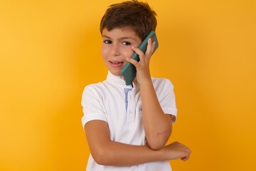 Portrait of a smiling little cute boy kid wearing white t-shirt against yellow wall talking on mobile phone. Business, confidence and communication concept.