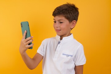 Isolated shot of pleased cheerful little cute boy kid wearing white t-shirt against yellow wall, makes selfie with mobile phone. People, technology and leisure concept