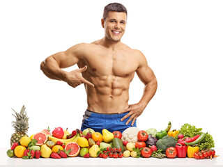 Fototapeta premium Bodybuilder posing with a pile of fruits and vegetables and pointing at his abs