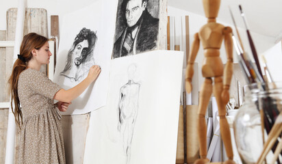 Indoor shot of young female artist standing in front of some sketches and drawing on it in bright white studio wearing 
bohemian chic clothing with painting tools in the foreground focus on background