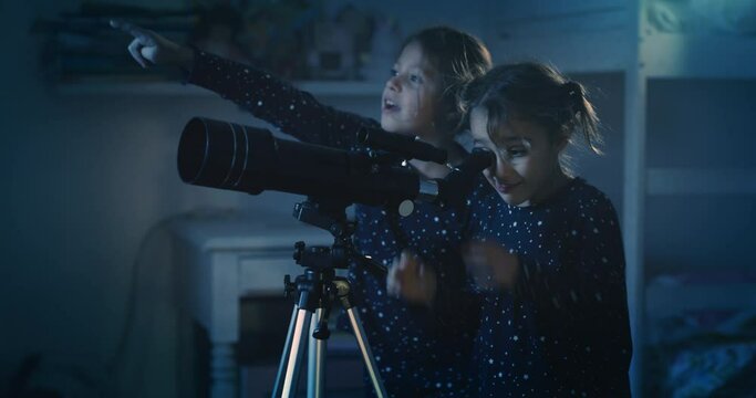 Cinematic night shot of two happy little girls sisters wearing starry pajamas are having fun to watching through telescope at falling stars in the dark sky. Concept: exploration, education, childhood