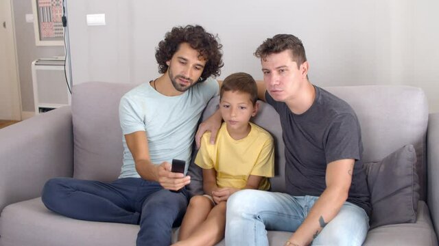 Two focused dads and son studying remote control for new TV, sitting in living room together, pressing buttons and talking. Family and home entertainment concept.