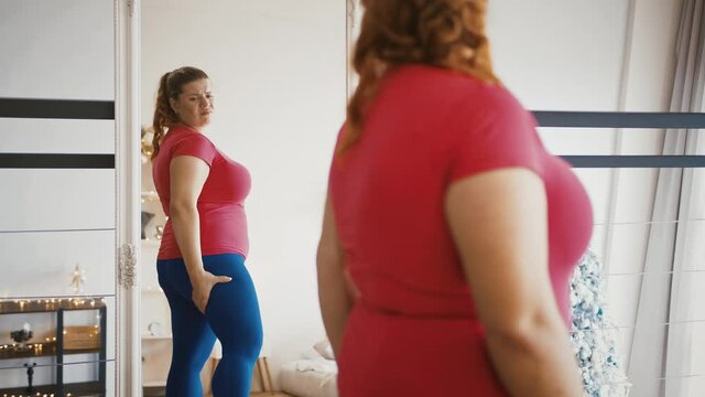 Young overweight woman feeling sad because of excess weight, looking at herself at mirror