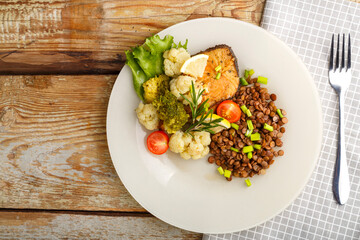 Baked fish with broccoli and lentils in a white plate with rosemary and lemon on a napkin next to a fork.