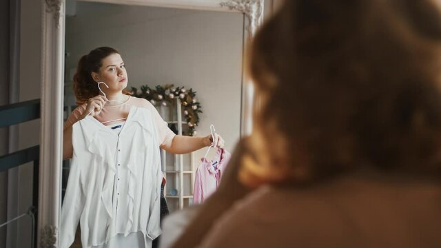 Oversize lady choosing blouse in dressing room, looking at mirror, plus size fashion