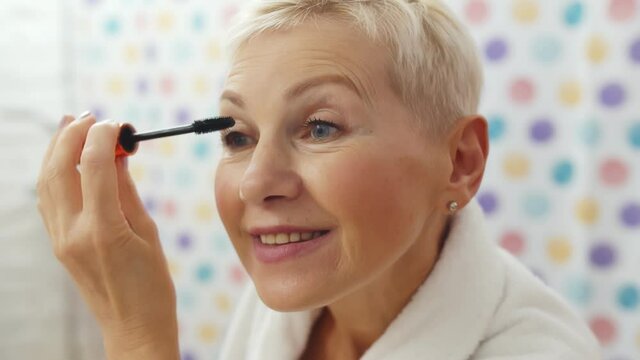 Mature woman putting mascara looking in mirror getting ready in morning
