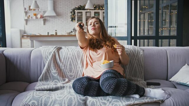 Young overweight lady sitting on sofa at home, watching tv and eating junk chips