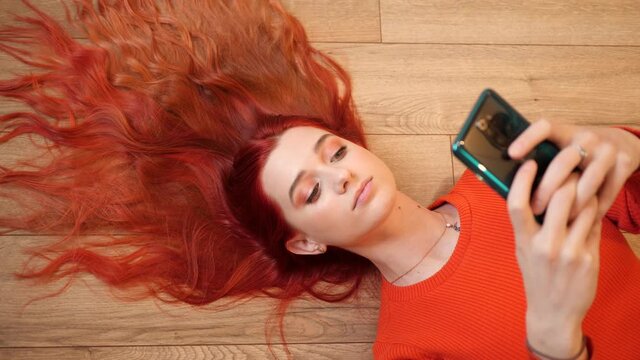 Redhead young pretty woman lie on a floor hair spreaded like a crown watch hold smartphone in social media