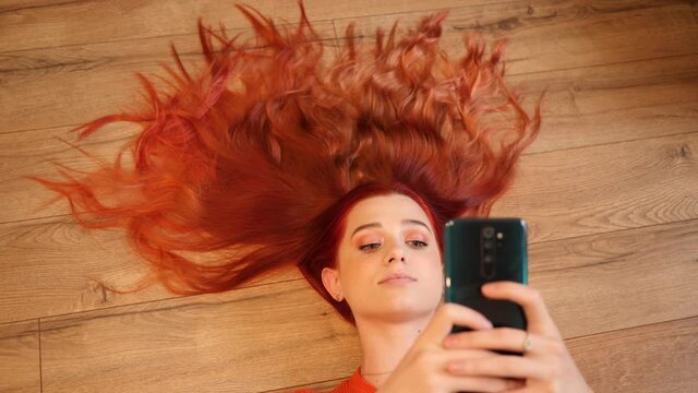Redhead young pretty woman lie on a floor hair spreaded like a crown watch hold smartphone in social media