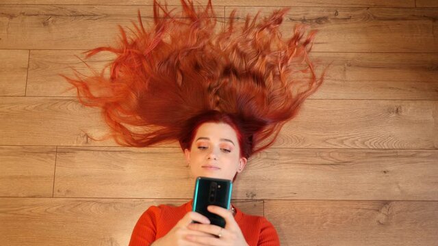 Redhead young pretty woman lie on a floor hair spreaded like a crown watch hold smartphone in social media
