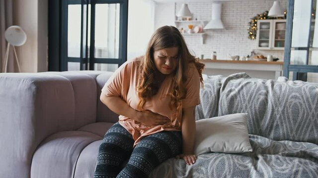 Young overweight woman suffering from abdominal pain, period cramps, sitting on couch at home