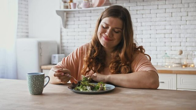 Happy overweight woman eating salad with great pleasure, enjoying healthy food at kitchen interior