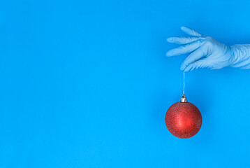 Hand in medical glove holds red christmas ball on blue background with copy space.