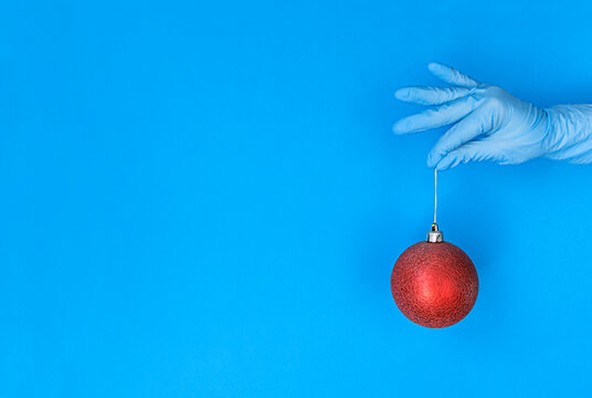Hand in medical glove holds red christmas ball on blue background with copy space.