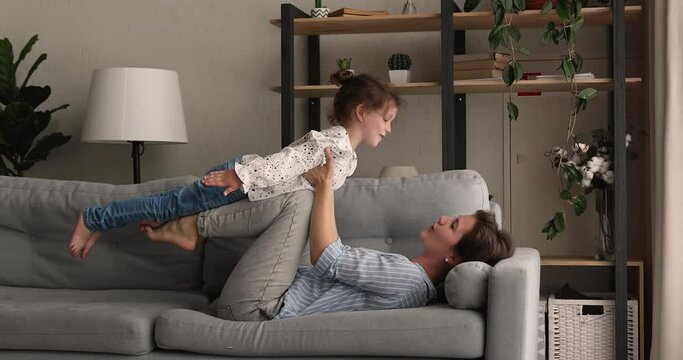 Side view young mother lying on sofa lifts up small cute daughter holding her on raised arms while kid imagines herself like plane, family dreaming about vacation travel and holidays, playtime concept