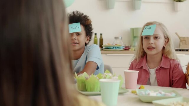 Medium over shoulder arc of multiethnic school children sitting at table on birthday party playing Who Am I Game. Boys and girls having sticky notes on their foreheads, talking and smiling