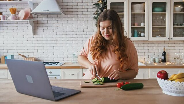 Overweight woman cooking salad at kitchen, watching video recipe on laptop online, cutting cucumber