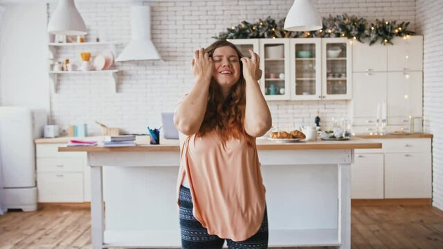 Body positive and energy. Active overweight woman enjoying dynamic music in headphones and dancing at kitchen