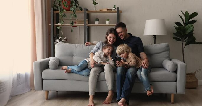 Happy family with little kids sit on couch in comfortable warm living room spend free time using smartphone apps. Educate children using modern tech, gadget overuse bad habit, have fun at home concept