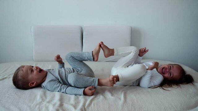 family, innocence, infant concepts - Two smiling children do leg exercises laying on white bed. Siblings little boy and girl brother and sister have fun and laughing, happy kids on quarantine at home.