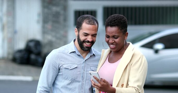 Interracial couple laughing in street holding cellphone. Candid mixed couple relationship looking at smartphone. Real life laugh and smile