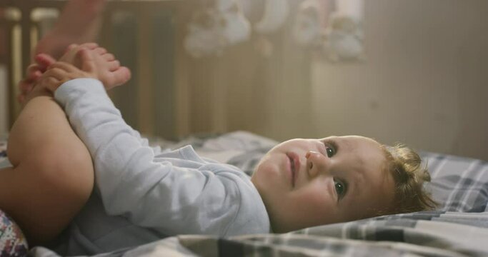 Cinematic close up shot of young happy mother is tickling and playing with her toddler baby boy in a nursery woke up in a morning. Concept of love, parenthood, childhood, life, maternity, motherhood.