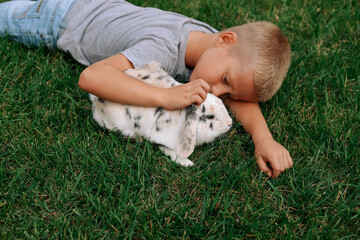 A happy blond boy lies on the green grass with his pet. A lop-eared tricolour rabbit.