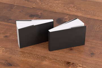 Two softcover or paperback horizontal or landscape black mockup books standing on the wooden background. Blank front and back cover