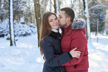 Fototapeta premium Young couple in love, enjoying winter weather outside in park forest. Brunette man in red jacket kissing woman in black, hugging, looking at each other. Christmas and new year holidays. Romantic date.