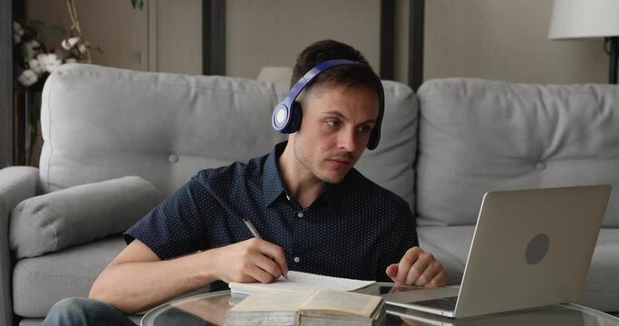 Focused guy sit in living room wear headphones listens audio course on laptop writing notes in copybook. Self education, making research using internet, study and e-learning using modern tech concept
