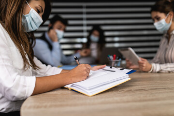 Group of four business people working together in their office on important project. They are wearing face protective masks, talking and analyzing their business plan together.  Covid-19 concept.
