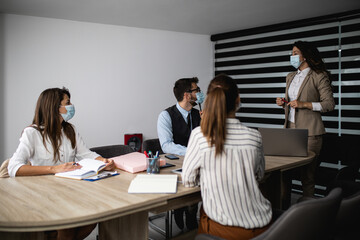 Group of four business people working together in their office on important project. They are wearing face protective masks, talking and analyzing their business plan together.  Covid-19 concept.