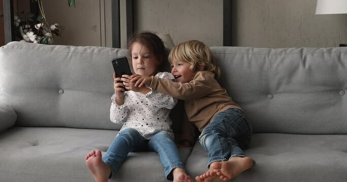 Cute little girl holding smartphone play games having fun with preschool brother sit on couch at home. Parental control app software, internet safety for children while kids spend time online concept