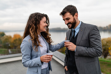 Male and female business colleagues standing on the roof of modern building during work timeout. They are drinking coffee, smiling and talking. Beautiful river landscape in the background.