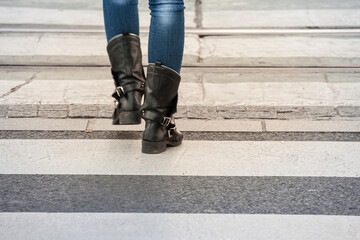 Feet of a woman with boots walking on the white strips of a pedestrian zebra crossing, or crosswalk, to pass a street and road used by cars, with a movement blur on her feet