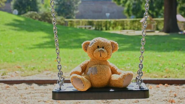 Cute little teddy bear standing alone on a swing in empty park on a beautiful sunny day.