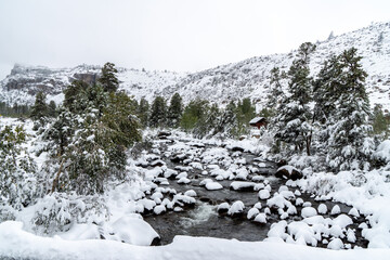 A river, stream,  with rocks and bank covered with fresh clean deep snow with  a cabin and trees and mountains in the background, Popo Agie River, Lander, Wyoming © Ron