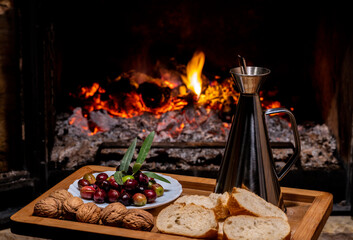 Aluminium oil cruet with flavoured olive oil on a wooden tray accompanied by raw olives, bread and nuts, over a rustic fire