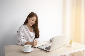 An Asian business woman sitting at a desk in her office. A laptop and a coffee mug on the desk.