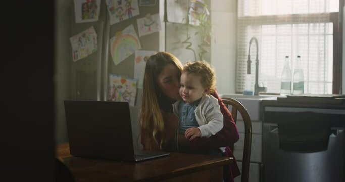 Cinematic shot of young happy mother and her smiling toddler baby boy having fun to make selfie or video technology call to family or friends with laptop after dinner in kitchen at home.